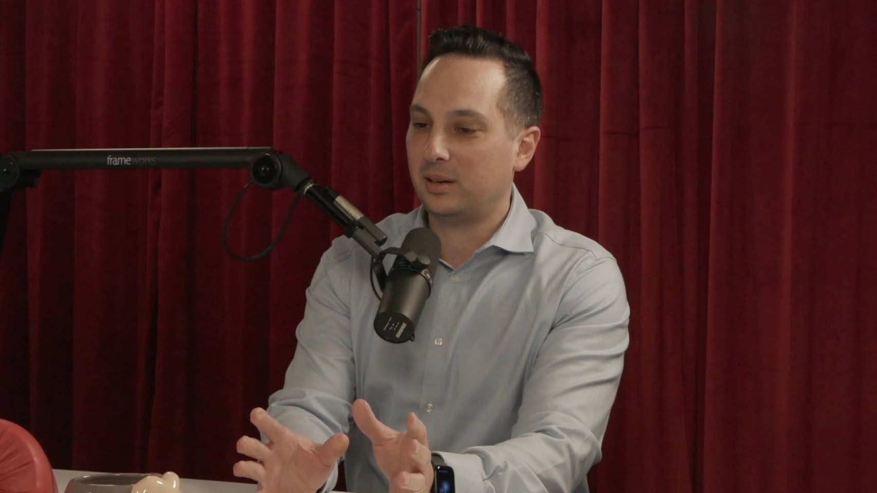 A man in grey shirt sitting at table with microphone.